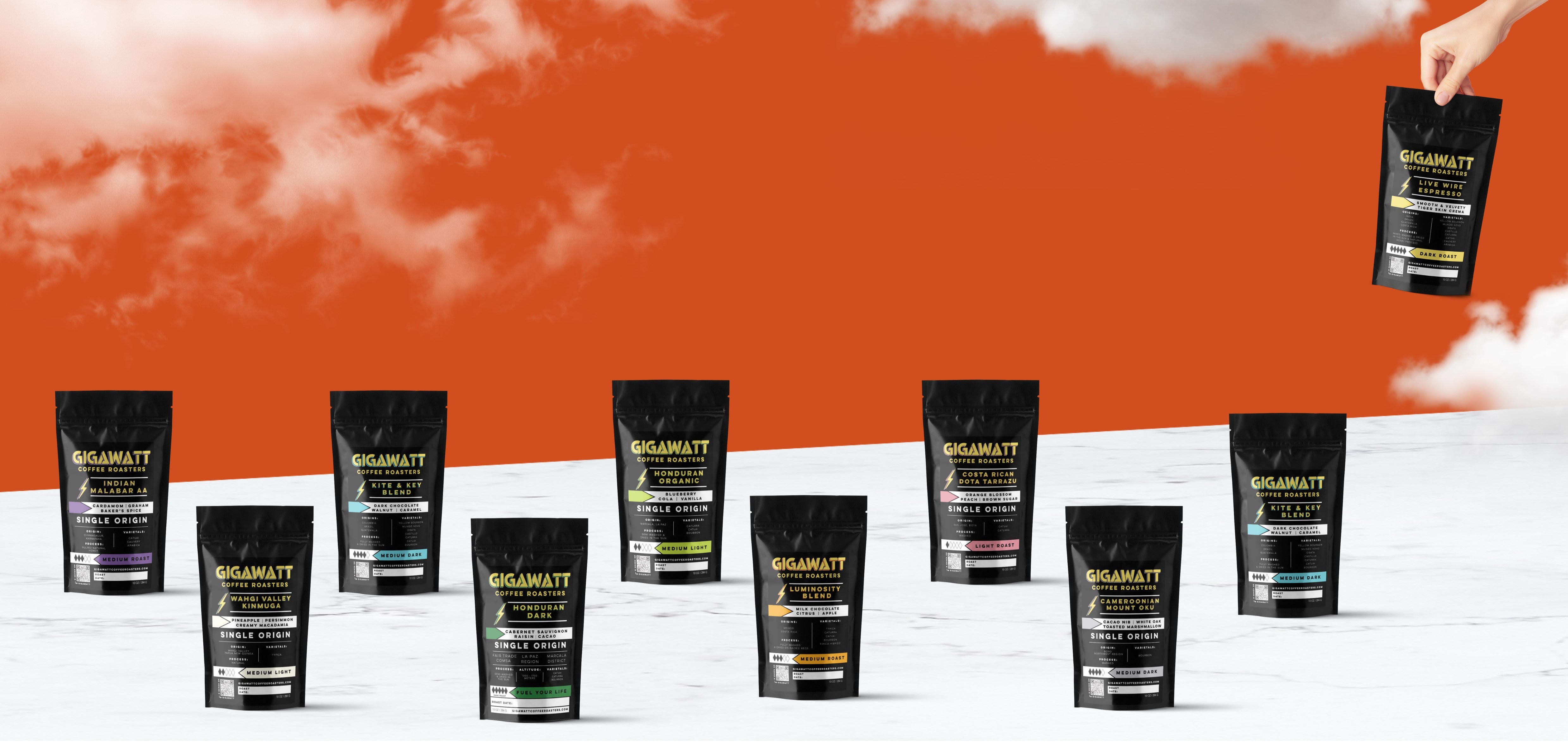 Gigawatt Coffee Roasters coffee sampler packs, featuring a variety of air-roasted coffee blends and single origin coffee like Indian Malabar AA, Kite & Key Blend, Costa Rican Tarrazu and more, displayed against a bold orange background with clouds.