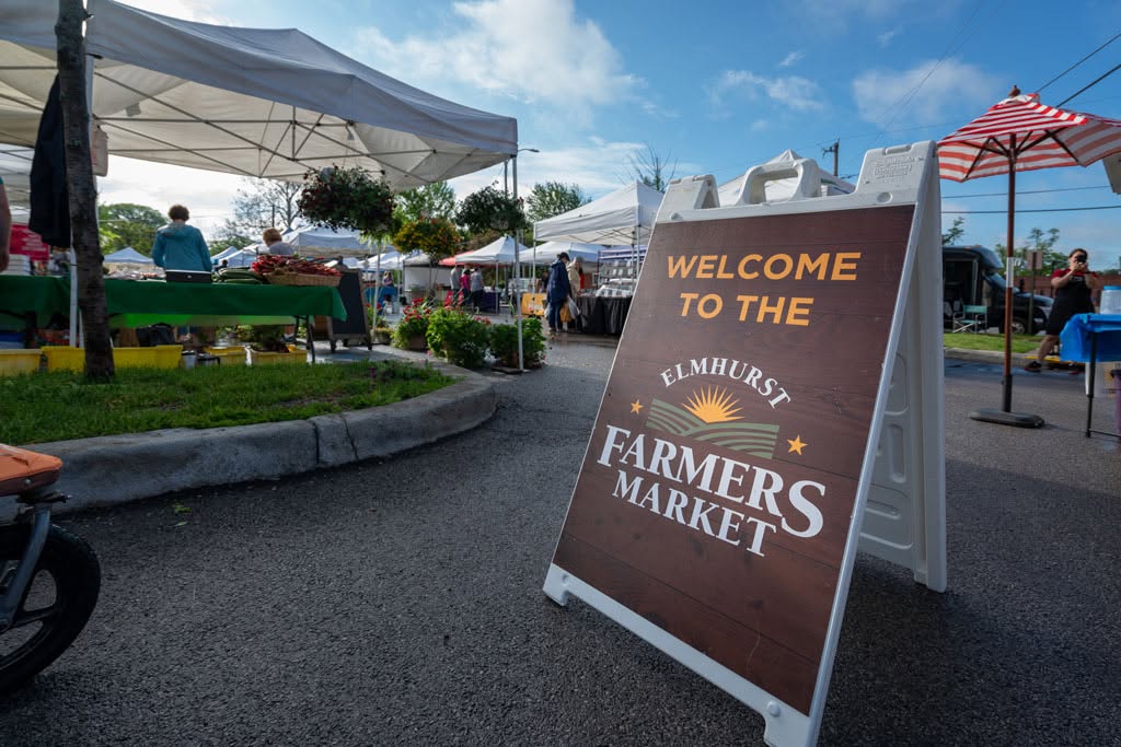 Welcome to the Elmhurst Farmers Market sign at the market entrance, with vendor tents and shoppers in the background