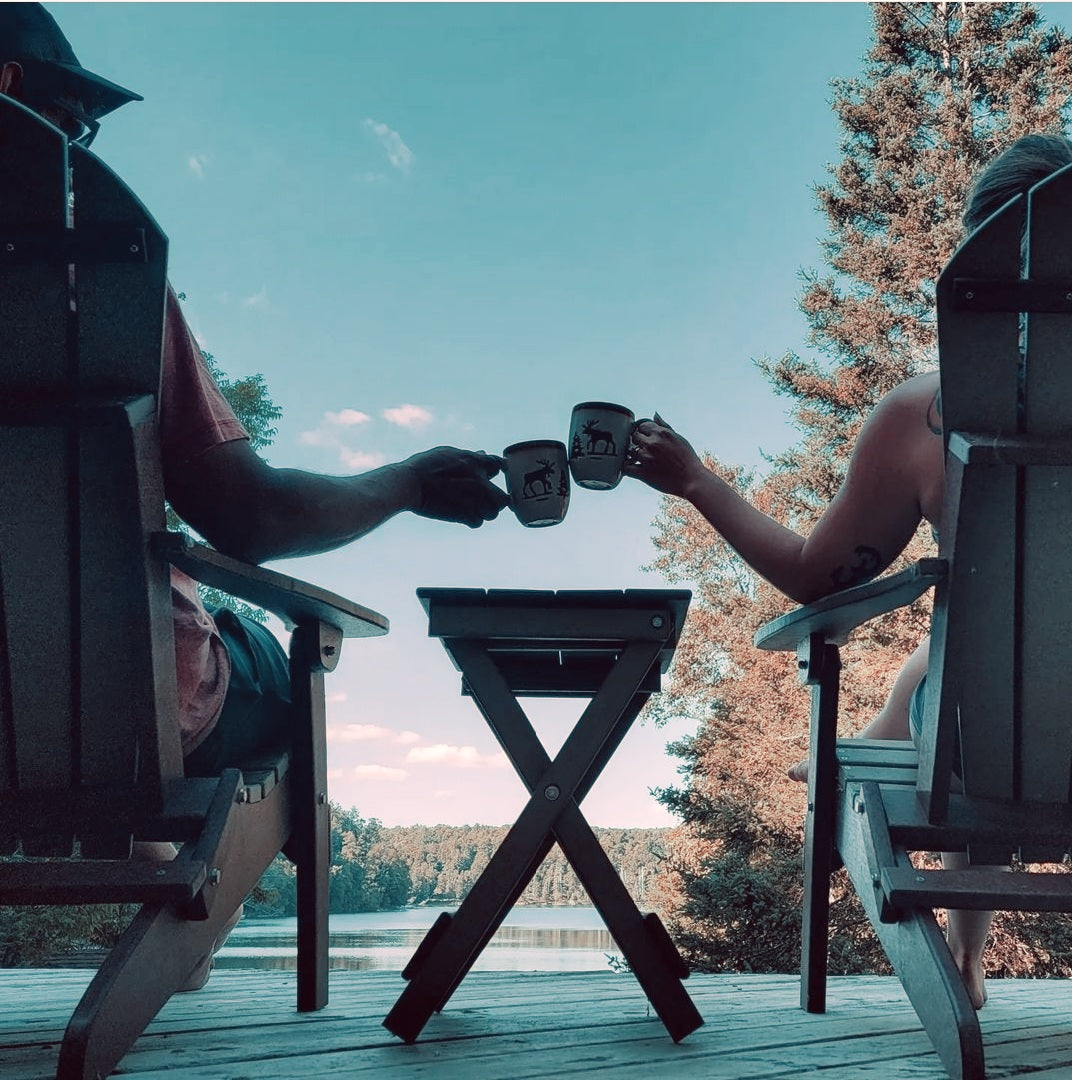 Two people enjoying Gigawatt Coffee on a lakeside deck, toasting mugs while sitting in Adirondack chairs, surrounded by serene nature and a clear blue sky.
